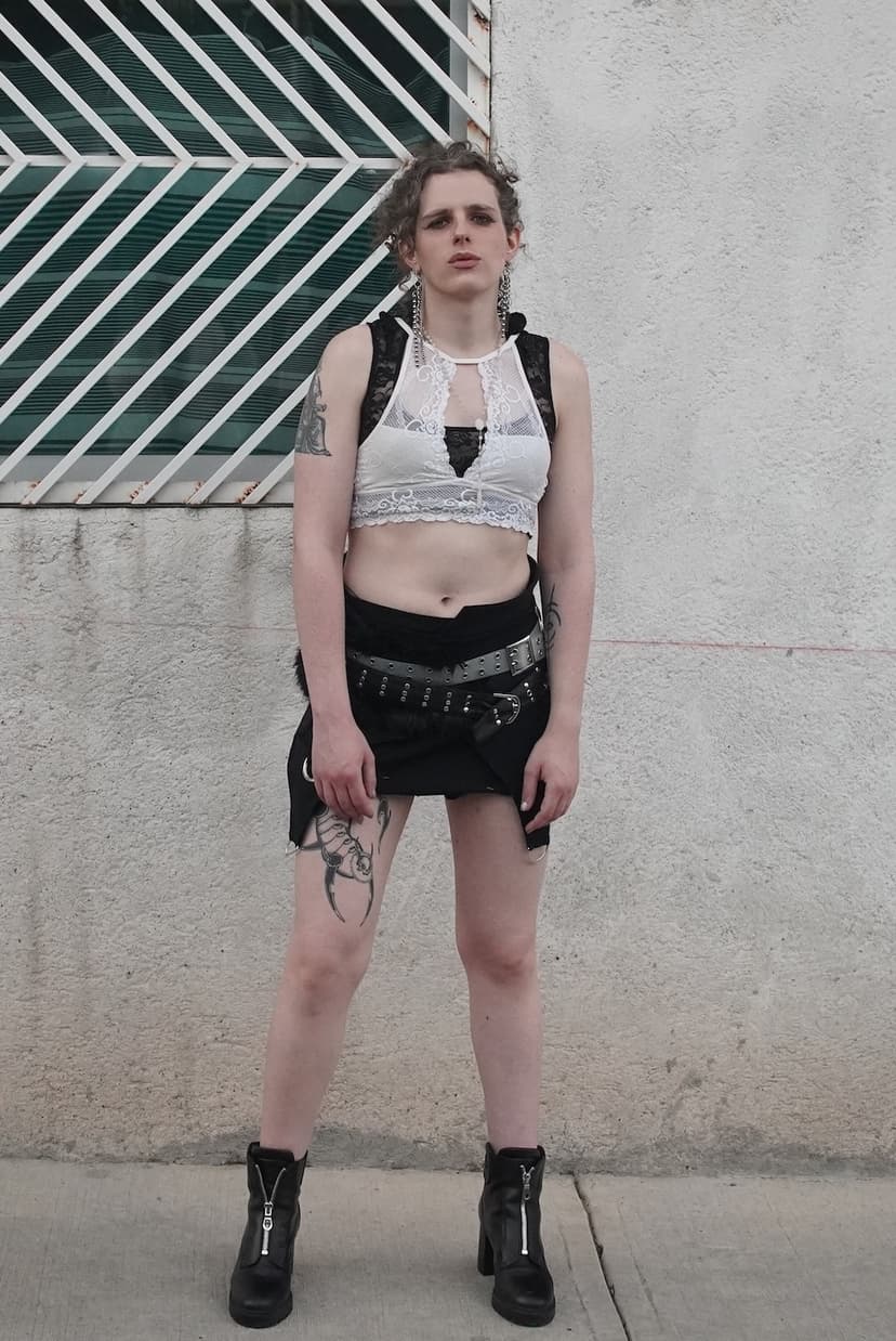 A woman in front of a white wall with a layered outfit of a crop top, a white bra, a skirt and some belts. She is wearing black boots with heels, long metal earrings, and has a big tattoo in her thigh.